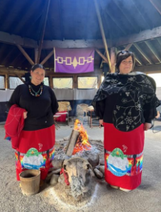 Inside a circular wooden lodge, two women stand beside a central fire. Both wear long red ribbon skirts with designs of a whale, turtle, rainbow, and floral bands, echoing the first artwork. They are dressed in black tops, one with a shawl, and behind them hangs a purple banner with the Haudenosaunee Hiawatha belt.