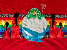 A textile artwork on a red background shows silhouettes of women in red dresses holding white feathers, standing against a rainbow and floral band. At the center, a large white circle contains a blue whale and a green turtle carrying a fire. Above is a constellation-like spider figure with a star.