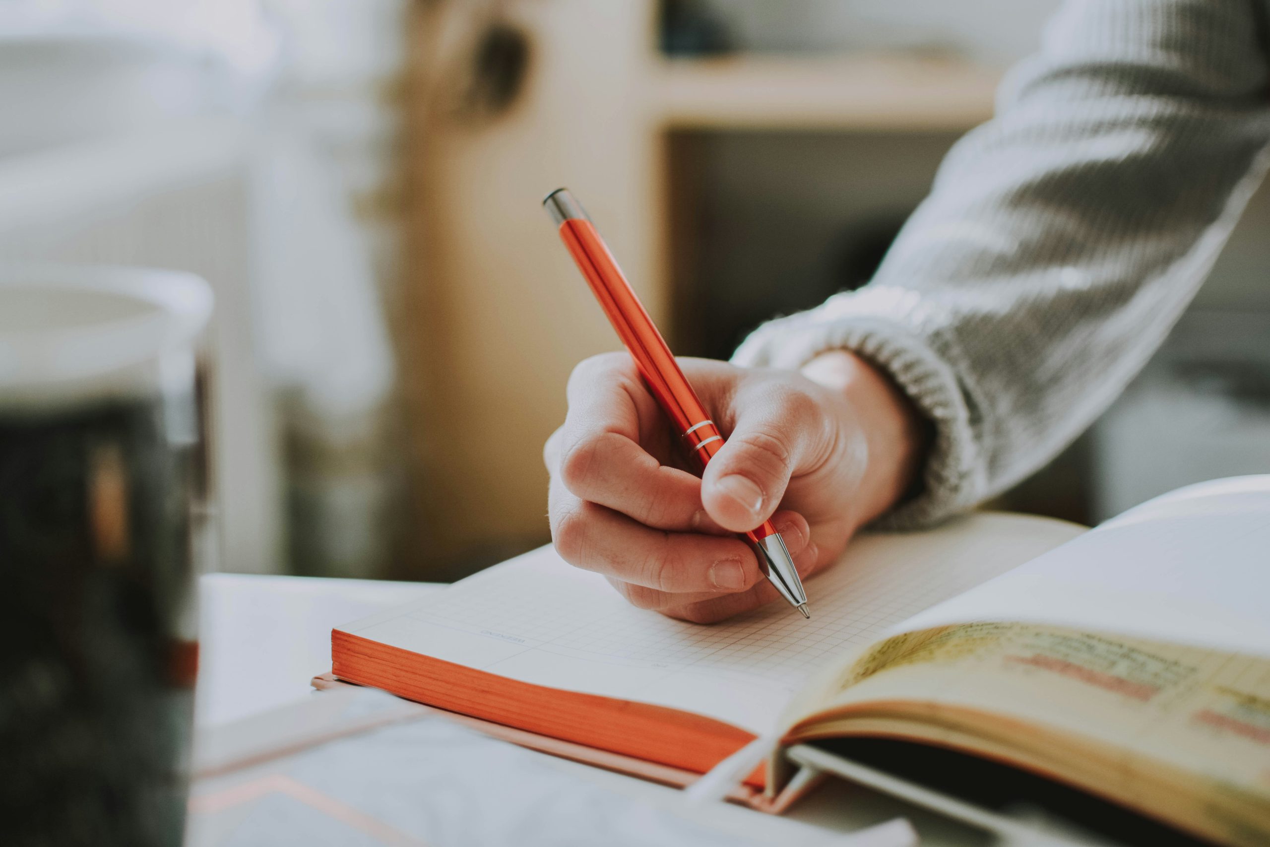 Photo of person holding on red pen while writing on book