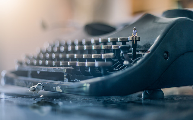Close up of a typewriter with a small person sitting on a key in the top row and a bird in the foreground
