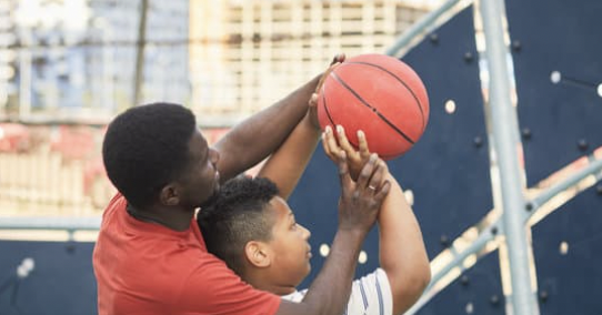 A coach teaches a student how to properly position hands for shooting a basketball by placing the dominant hand’s fingertips under the ball and the guiding hand on the side of the ball.
