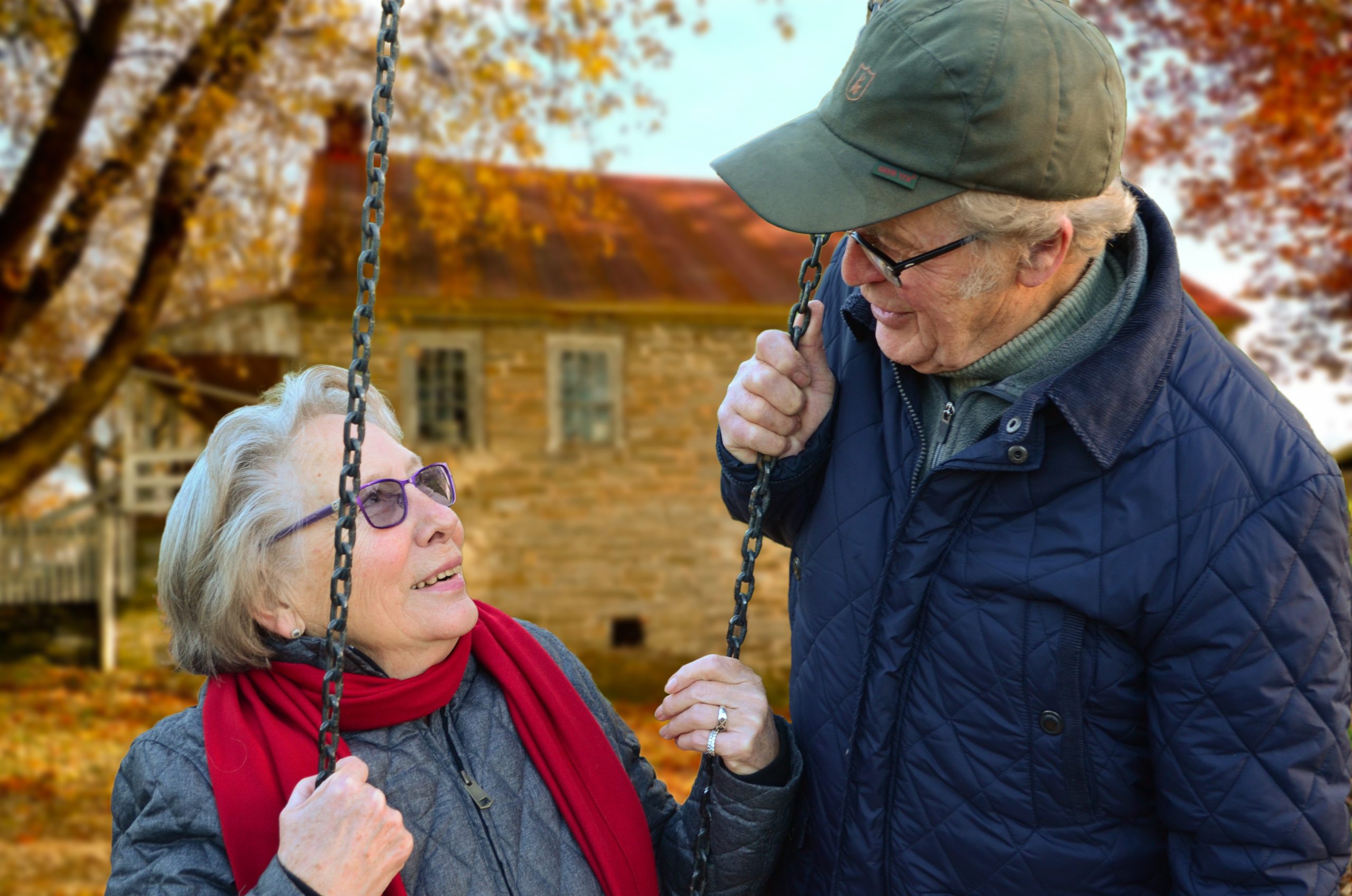 Picture for decorative purposes. Color photography of an elderly woman on a swing and an elderly man looking down at her. It is fall.