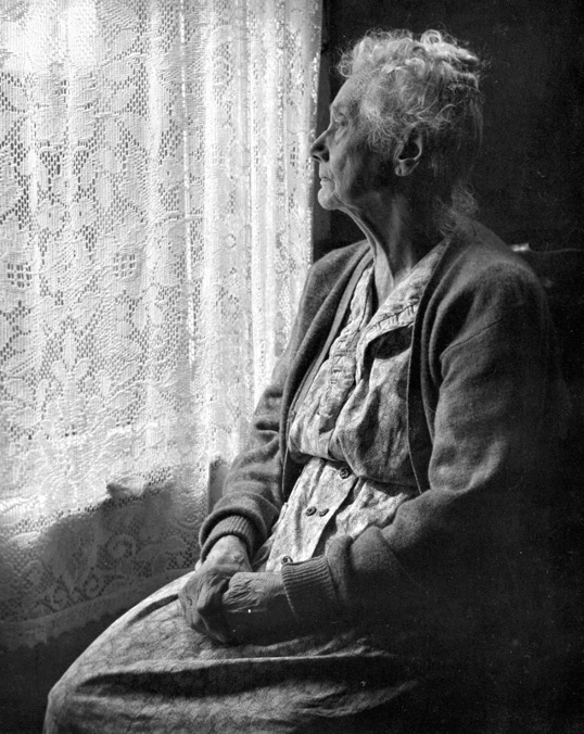 Black-and-white photo of a lonely elderly woman looking out of a curtained window.