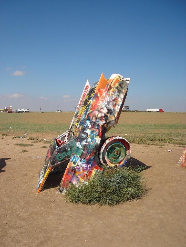 A single spray-painted Cadillac is pictured in front of a rolling field and blue sky.