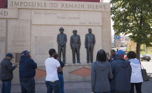 Photo of Clayton Jackson McGhie Memorial in Duluth, Minnesota. Top of memorial is end of statement, "Impossible to remain silent" above the names and bronze depictions of three men lynched in 1920: Elias Clayton, Elmer Jackson, and Isaac McGhie. Photo includes man talking with group of 7 people.