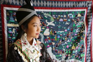 Photo of young Hmong woman wearing clothes representative of her family's clan. She is standing in front of a paj ntaub (story cloth) depicting migration journey.