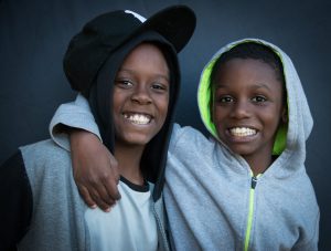 Two smiling adolescent boys. One has his arm around the other. One has sideways baseball hat and the other has sweatshirt hood.