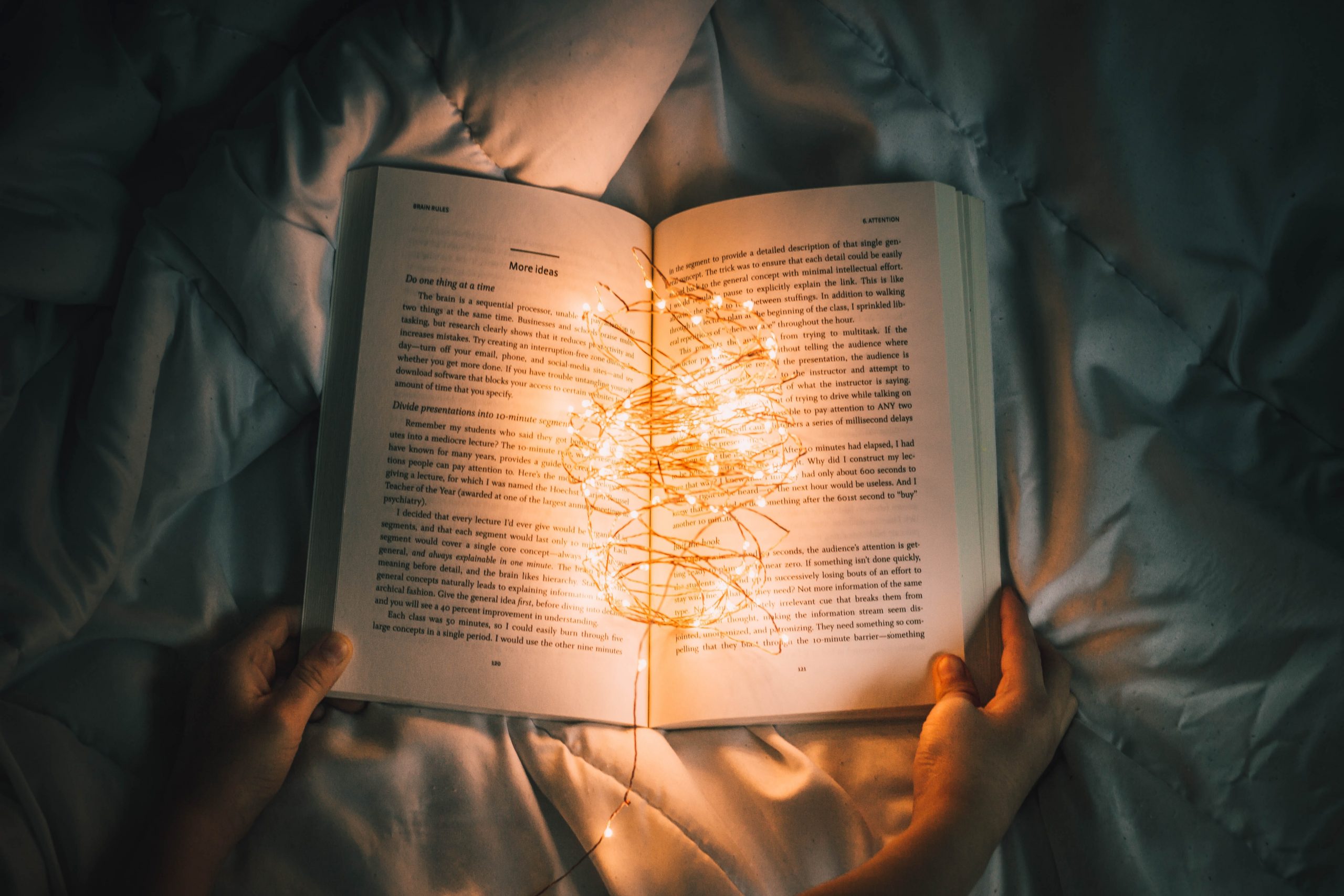 open book on the comforter of a bed with two hands holding it open. It has small string lights sitting in the center of the book, lighting up the words on the page, contrasting with the dim lighting surrounding it