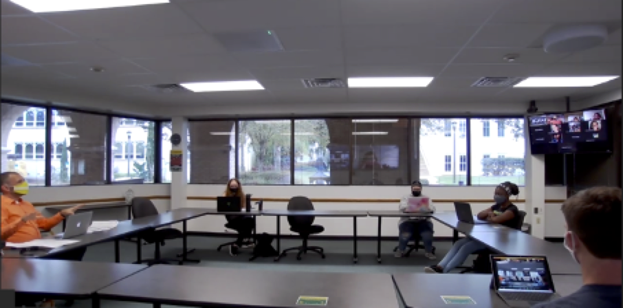 Classroom with a professor teaching his students. All the tables and chair are facing the center of the room forming a square and having all the chairs on the outside of the square. The professor is seated on the outside of the square with the students away from the podium that is out of frame. How the furniture is situated within the space encourages discussion and places the professor on equal ground with the students.