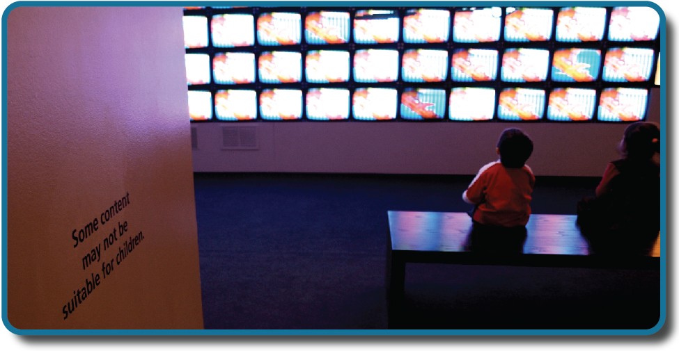 Children sit in front of a bank of television screens. A sign on the wall says, “Some content may not be suitable for children.