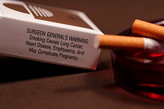 A photograph shows pack of cigarettes and cigarettes in an ashtray. The pack of cigarettes reads, “Surgeon general’s warning: smoking causes lung cancer, heart disease, emphysema, and may complicate pregnancy.