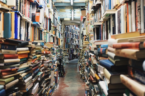 Looking down the aisle of library stacks with books overflowing onto floor in piles