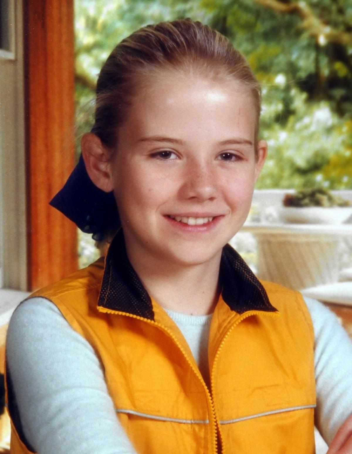 A photo of Elizabeth Smart as a little girl. She sits smiling at the camera in a yellow vest and long-sleeved white shirt.