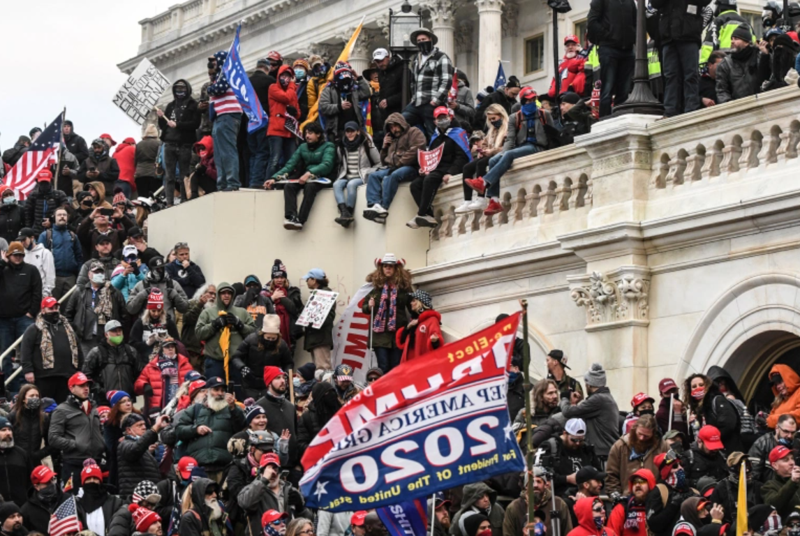 rioters sitting on and surrounding the US Capitol building and holding a Trump 2020 flag