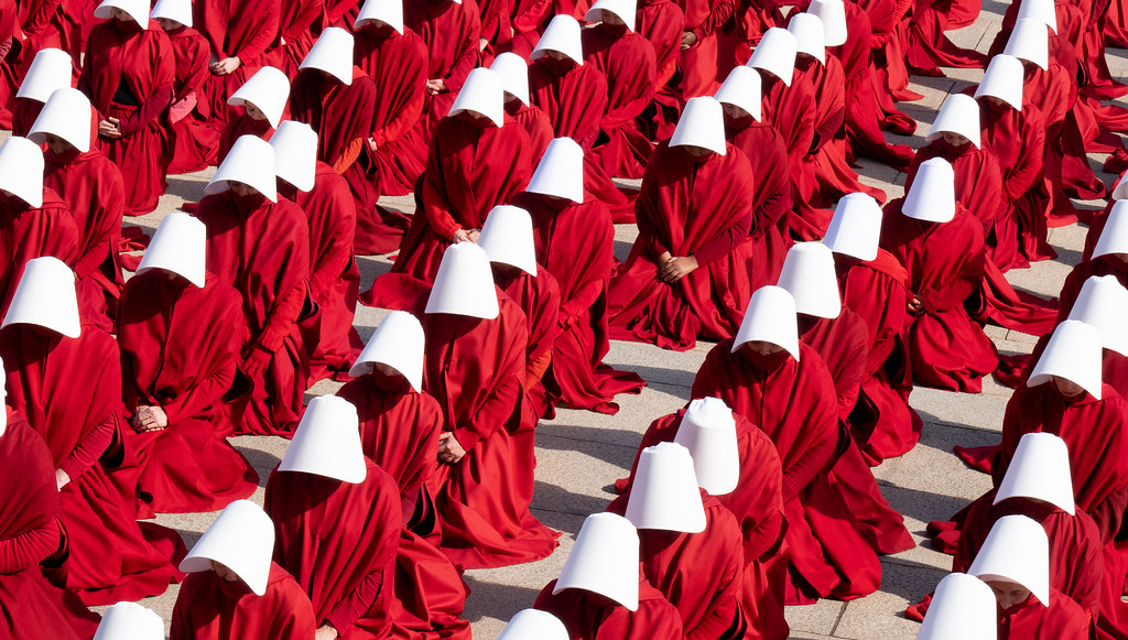 dozens of women kneeling in rows wearing white bonnets and red cloaks from the tv show The Handmaid's Tale