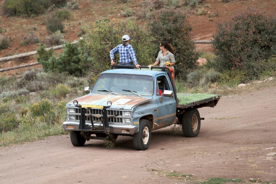 Photograph showing two people riding in the back of a flat-bed truck on a dirt road, holding onto a bar across the top of the truck