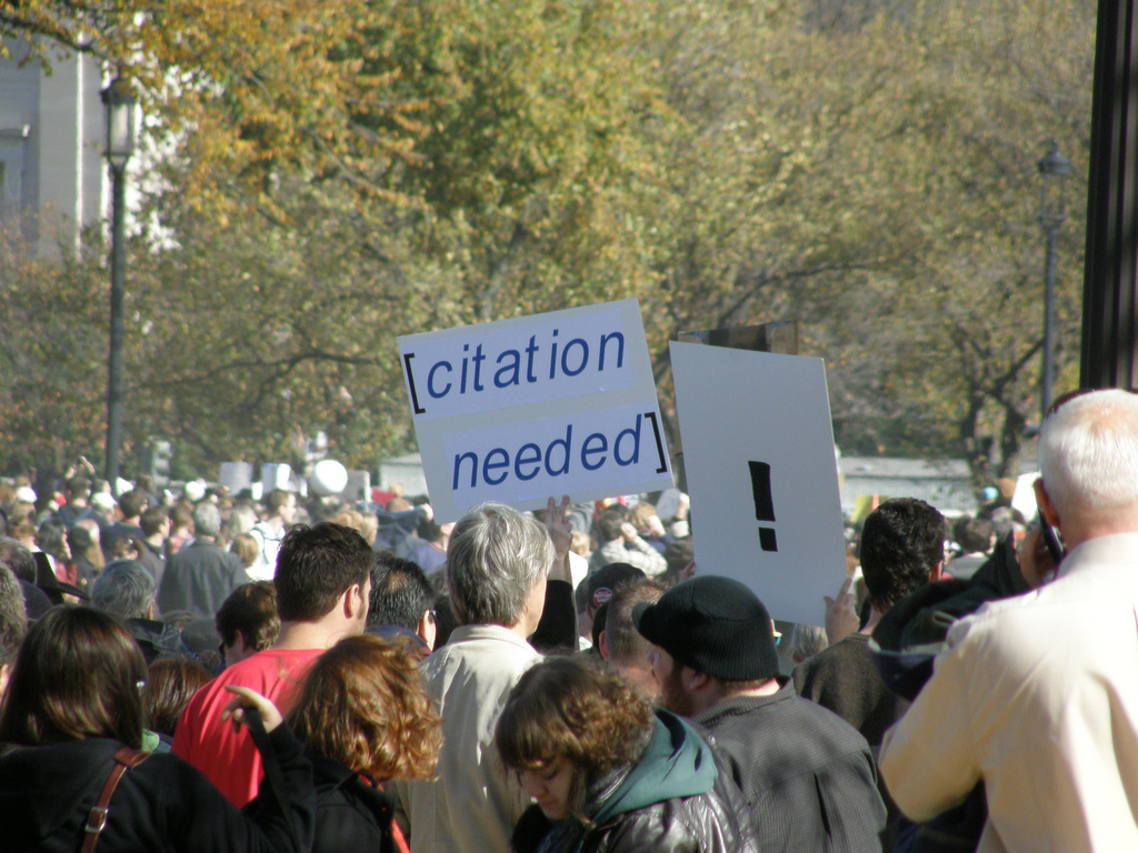 A crowd of people with one person carrying a placard with "!" on it and another with "citation needed" on it