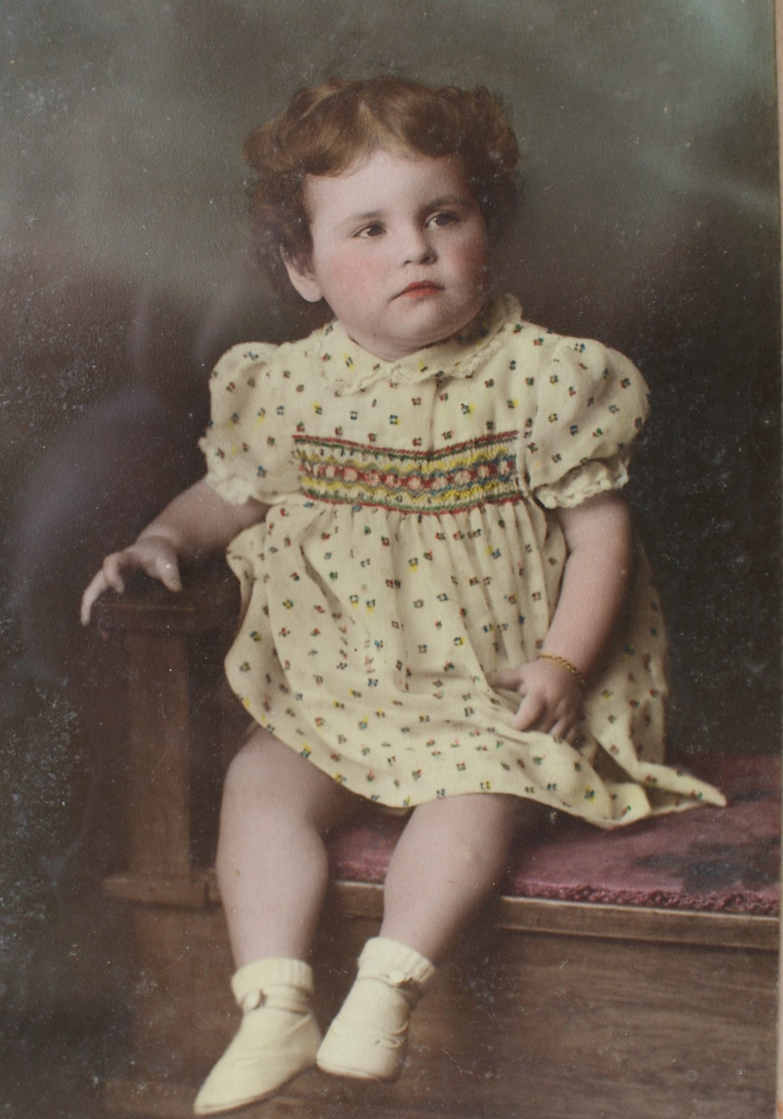 Lesley aged about 18 months seted on a stool in photographers studio. She is dressed in a smocked frock and looking off into the distance