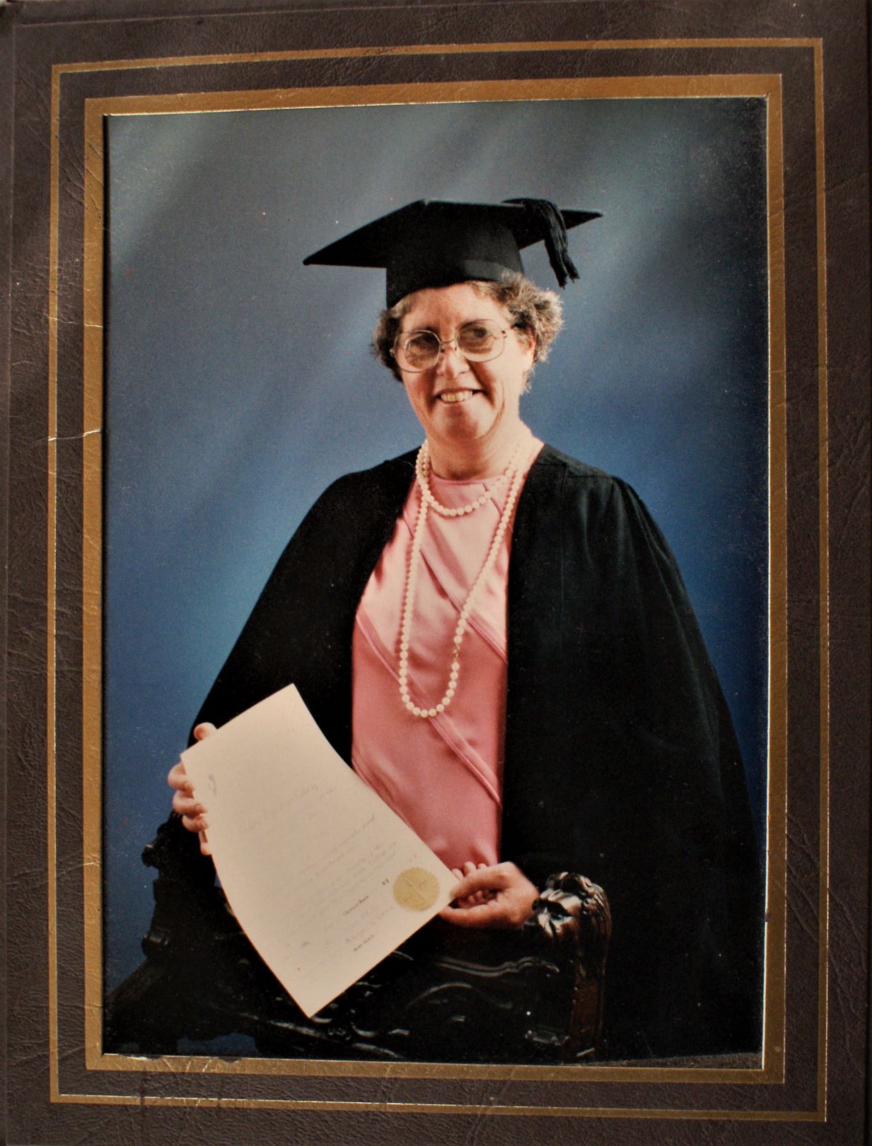 Studio shot of Lesley dressed in graduation gown and mortar board. Seated sand smiling, she holds her diploma