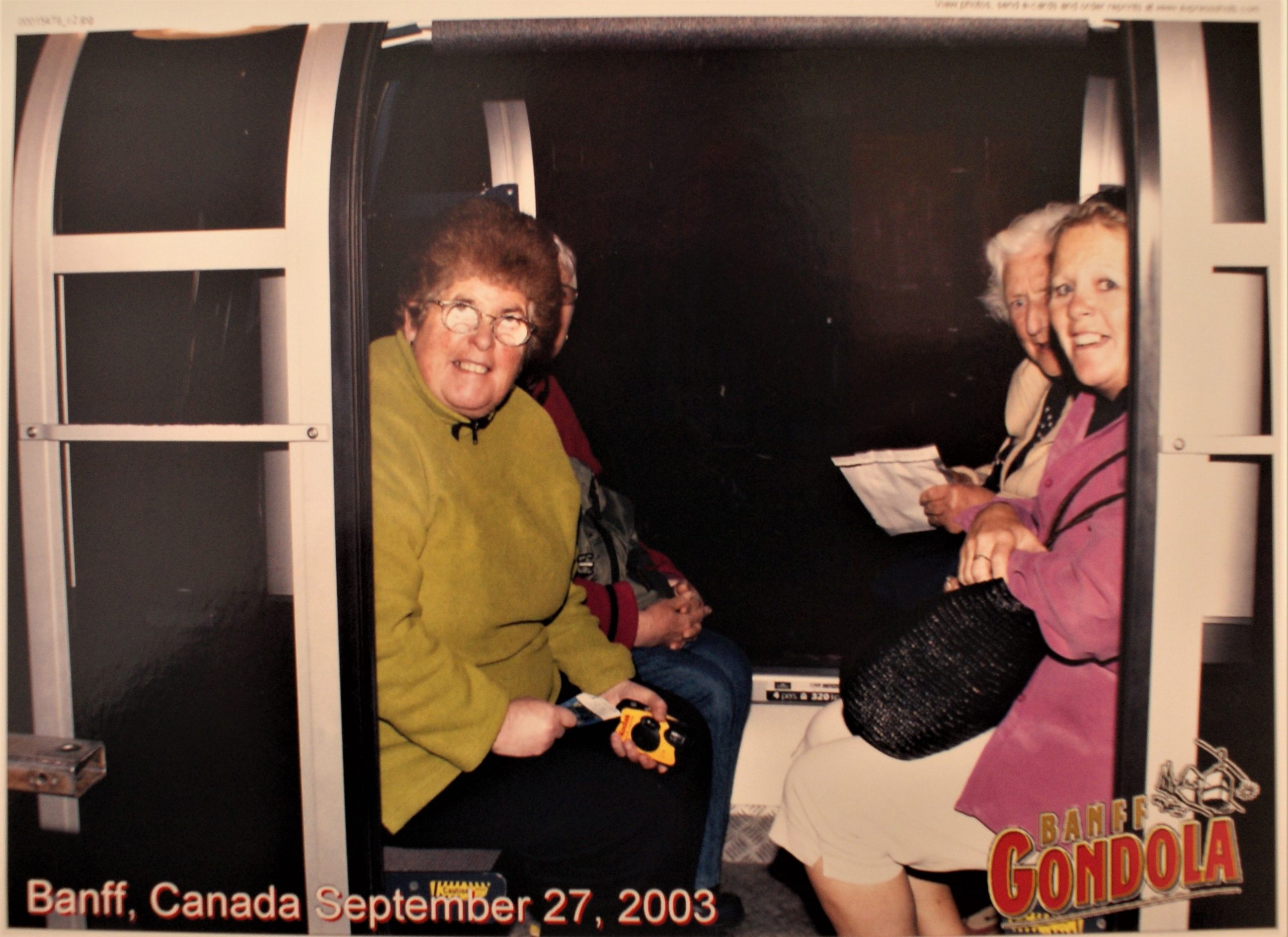 Lesley is looking out the open door of a gondola cart. Three other travellers are with her. The image is overprinted with the words Banff Gondola Banff, Canada September 27 2003