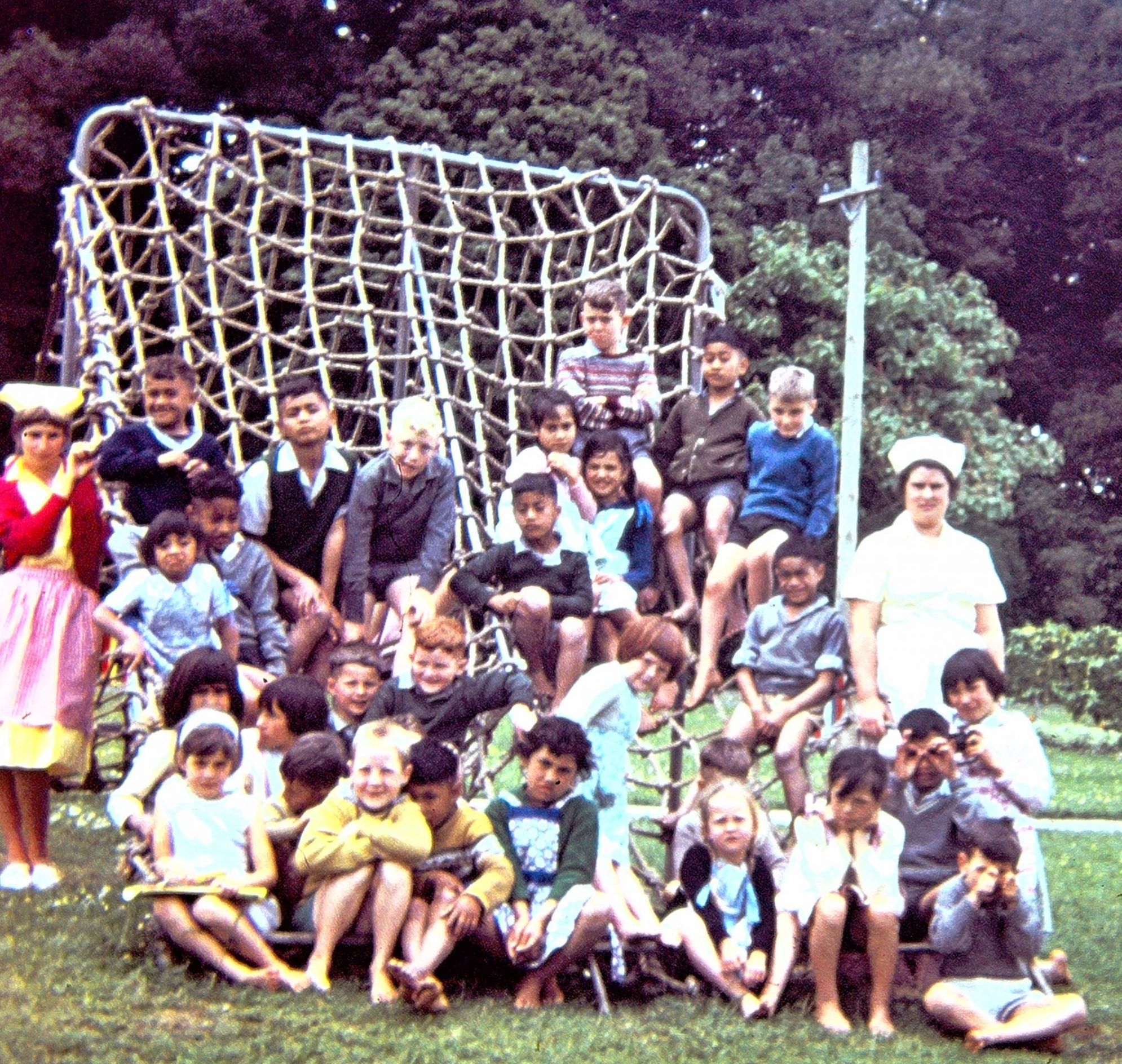 A group of children posed in front of a playground climbing frame with two staff in nurse-style uniforms
