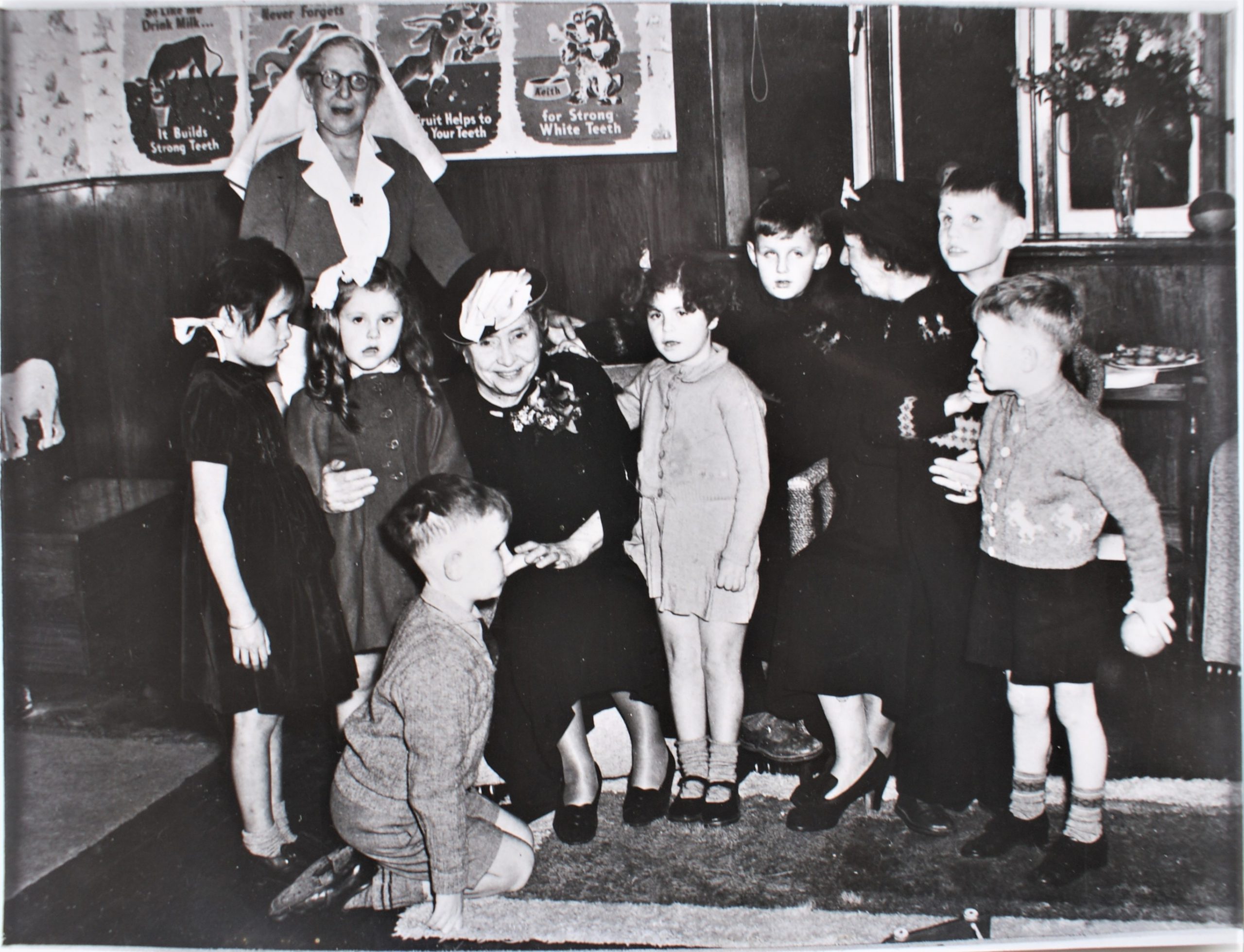 Helen Keller and a companion are sitting surrounded by seven children at the Sunrise home. At the back of the group is a woman in nurses uniform. On the wall behind are posters promoting dental health and a vase of flowers on the window sill.