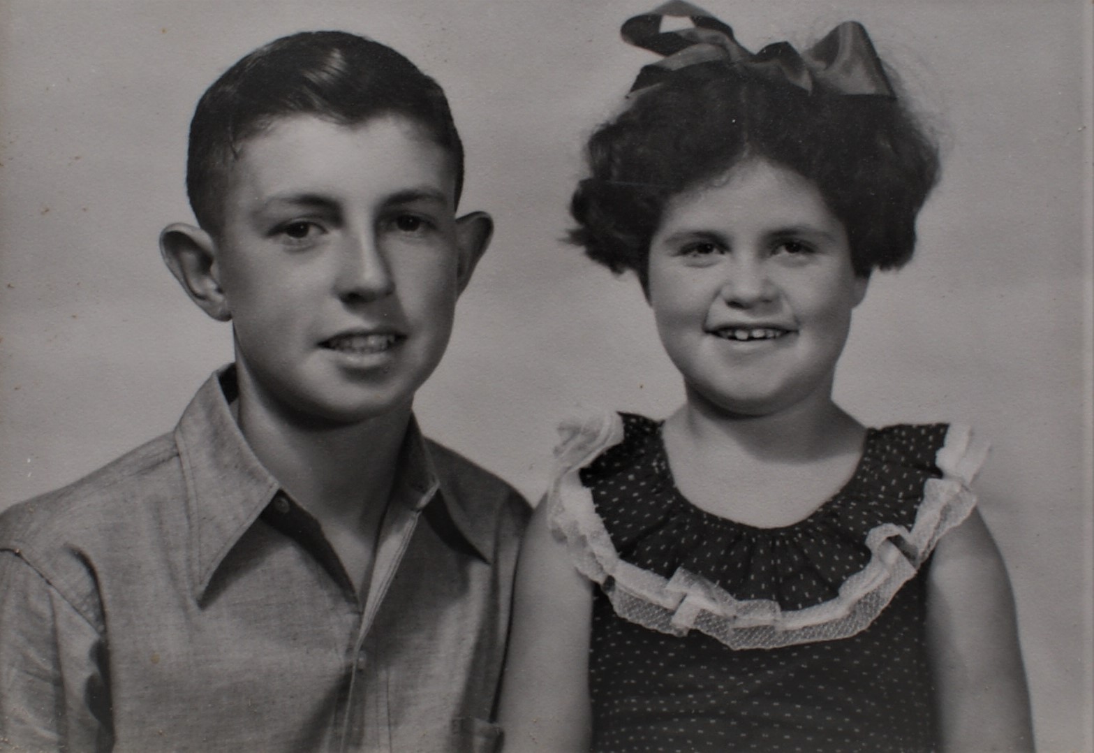 Studio shot of Lesley and her brother Ross. Both are looking straight at the cameraLesley wears a lack sleeveless dress with a frilled neckline and a bow in her hair