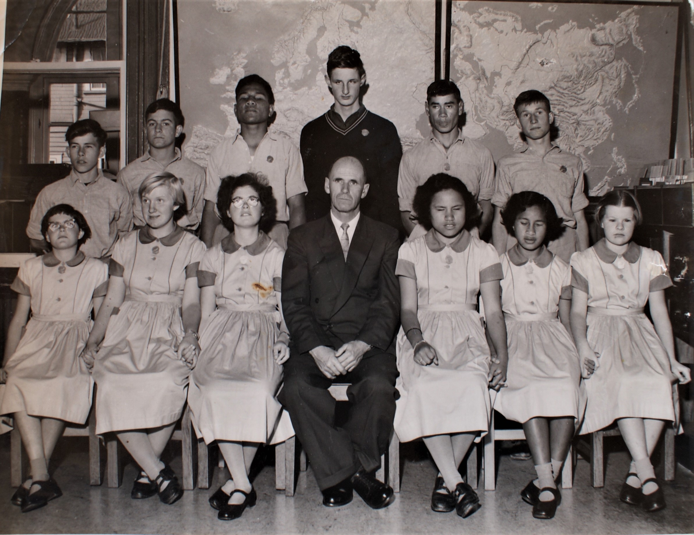 twelve students are seated in two rows for this formal photograph. The male teacher, wearing a dark suit is in the middle at the front. Lesley in school uniform and black shoes is sitting in the front row