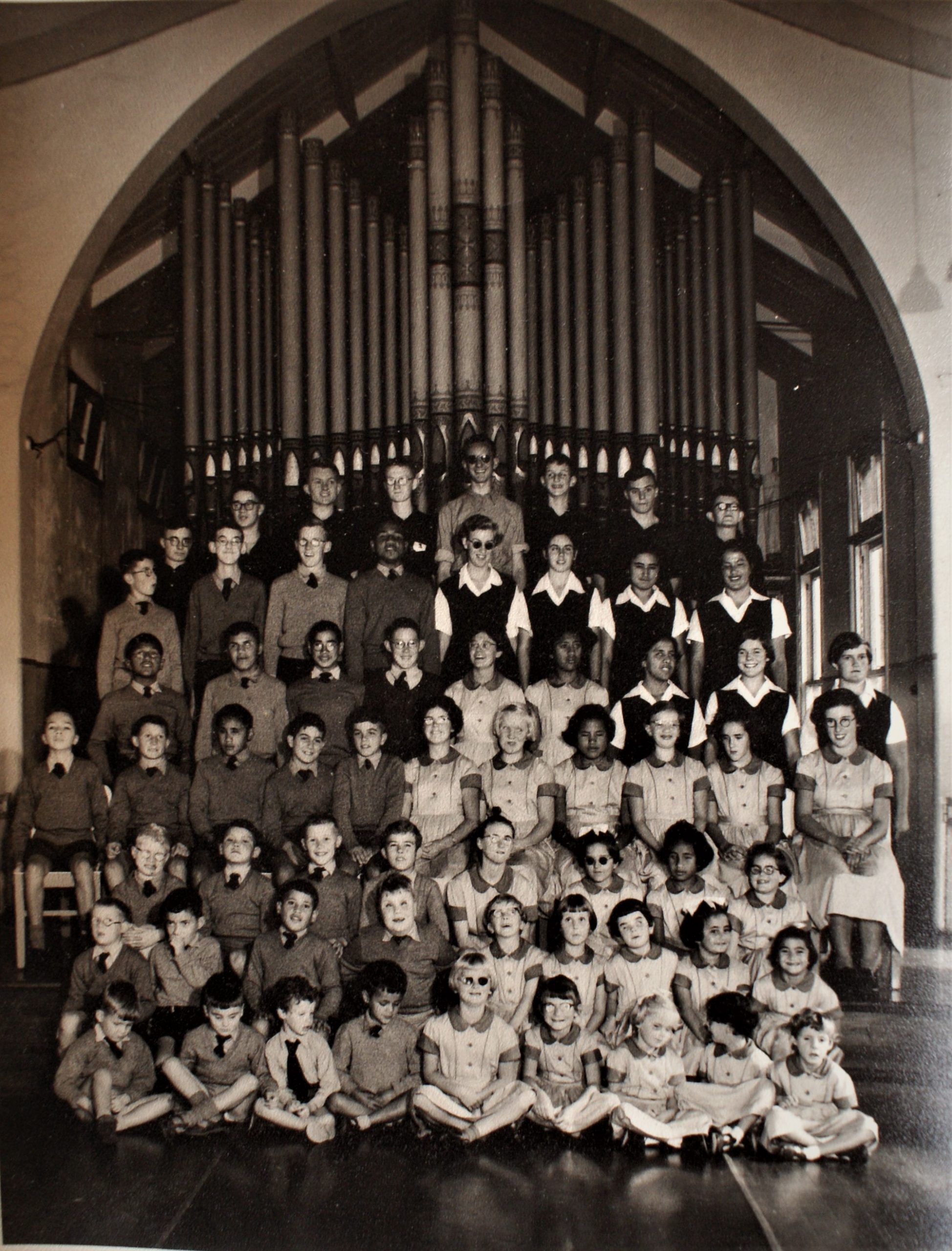 Seven rows of students are pictured in front of a set of organ pipes, possibly in a church. The older students st the back are standing. The youngest students are cross-legged on the floor in the front. Teenaged Lesley, wearing glasses and smiling is in the very centre of the group
