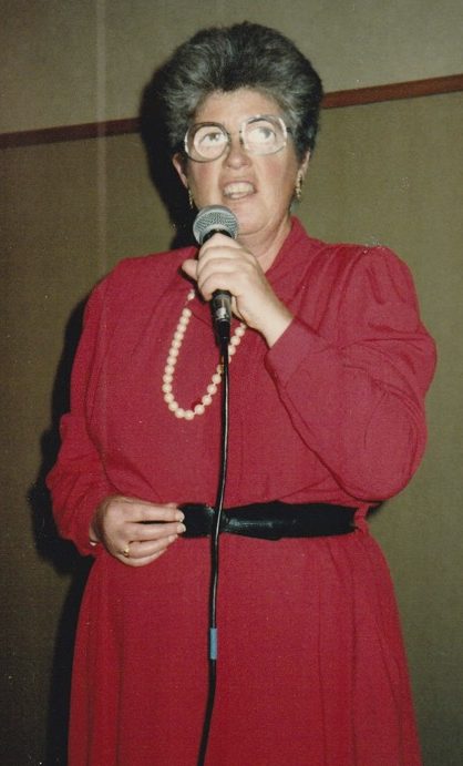 Lesley is standing speaking into a microphone. She is wearing a red dress with long pearl beads and a black belt