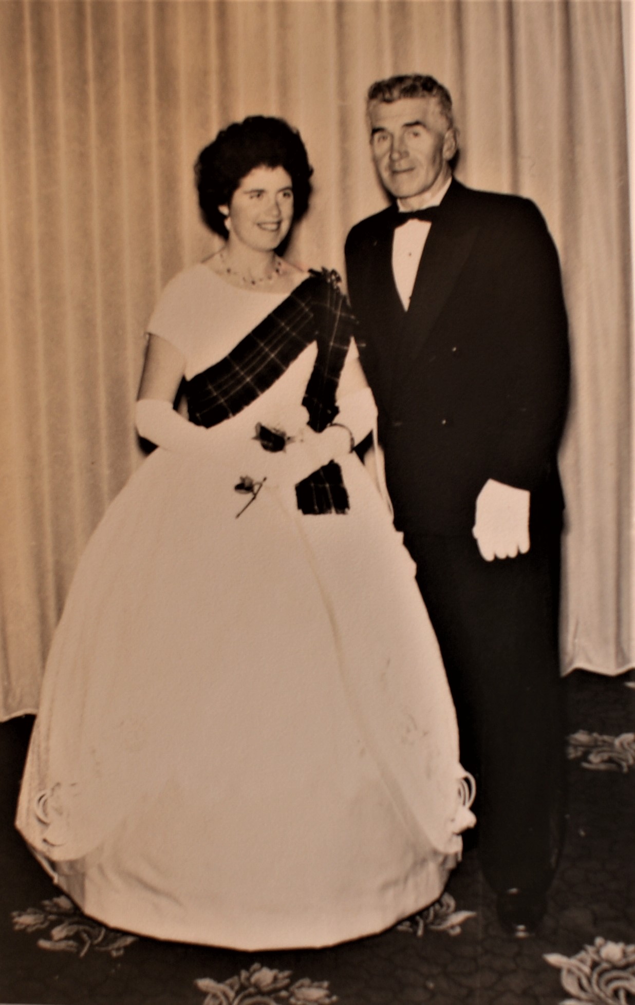 Debutante Lesley dressed in full length white gown with a tartan sash and long white gloves standing beside her besuited Father