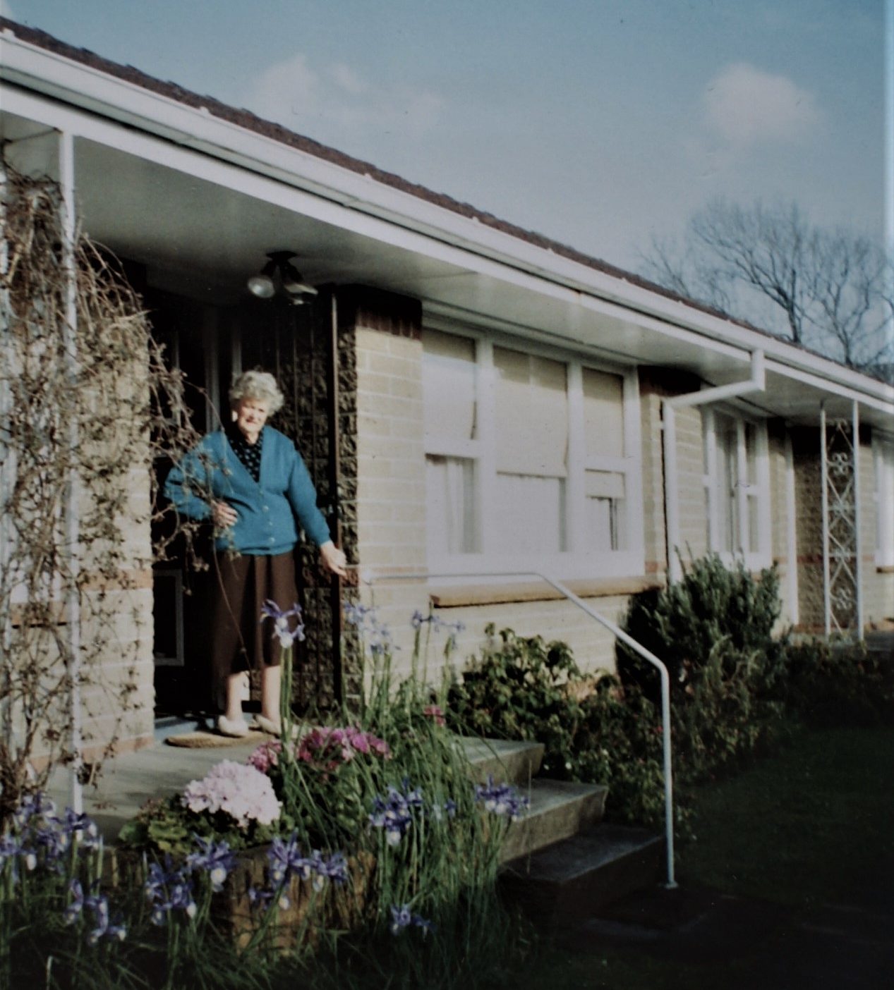 Lesleys Mother Dora stands in her slippers holding open the front door of a sand-coloured brick home unit. In front of the concrete terrace there are purple irises and pink flowers in bloom