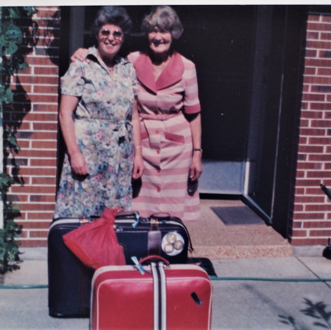Aunty Lucy has her arm over Lesleys shoulder. They are standing on Lucys front porch with Lesleys two suitcases, ready to go.