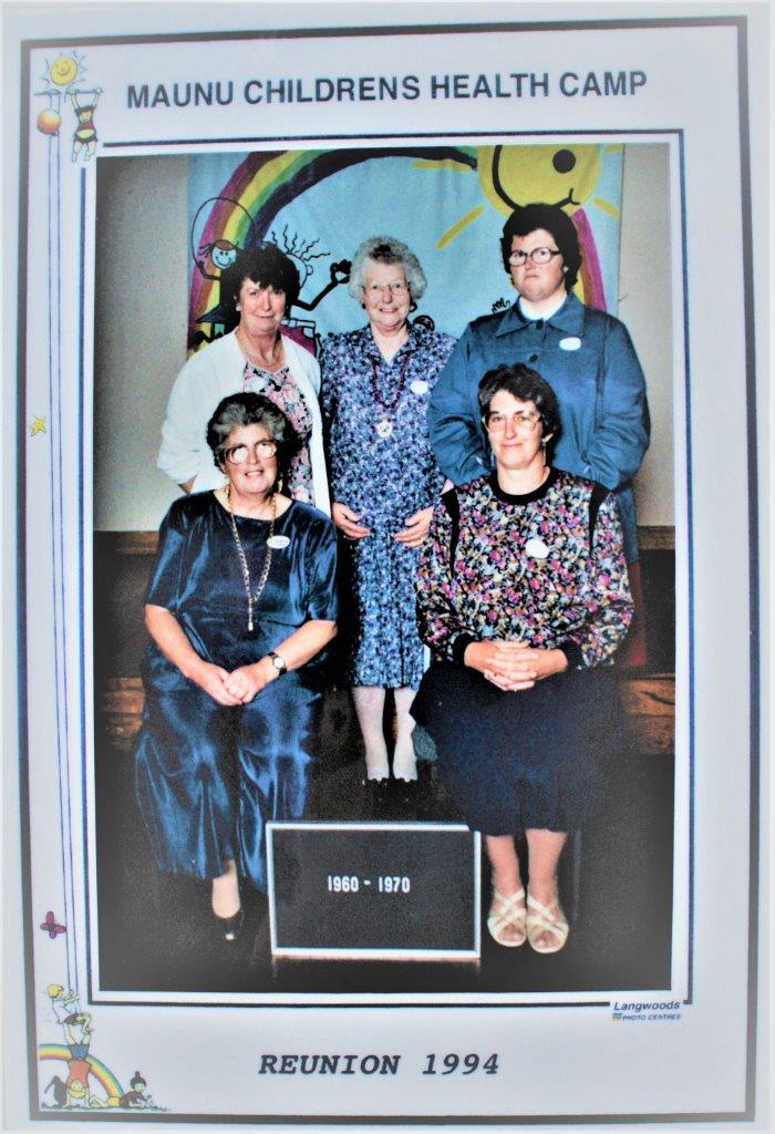 Three women are standing with Lesley and one other seated in front. Behind them is a wall hanging depicting a painted rainbow, stick figures of children playing and a smiling yellow sun. A board at the front of the group reads 1960-1970. The frame states Maunu Childrens Health Camp Reunion 1994.
