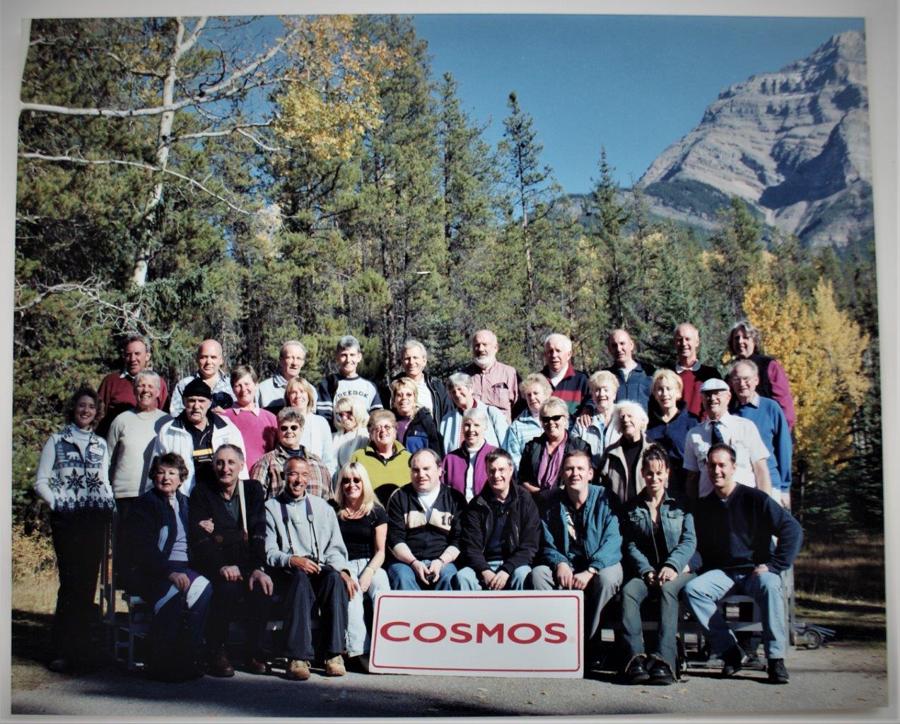 A tour group of about 35 people standing and sitting in four rows. Behind them is a blue sky, a rocky mountainside and forest trees. A sign in front reads COSMOS