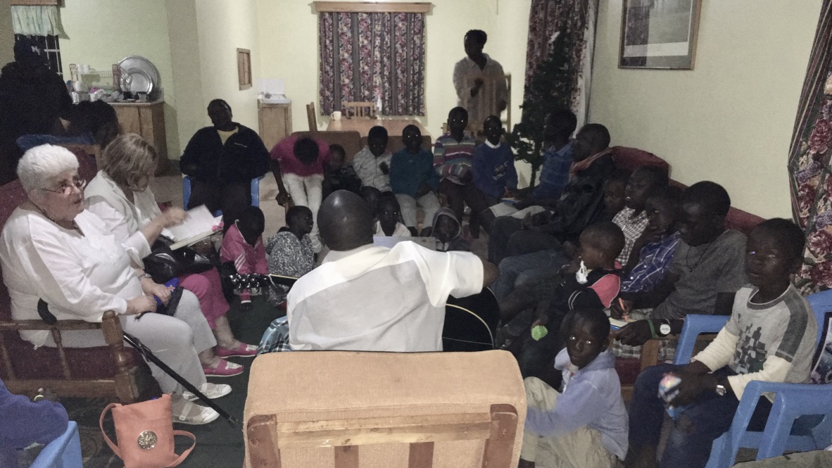 A Kenyan man with his back to the camera is sitting in an armchair with a guitar. On the floor and on chairs around him are seated 25 others, mainly children. Lesley holds in her lap a small NZ flag. and a friend from NZ are sitting in armchairs. Leslsy has