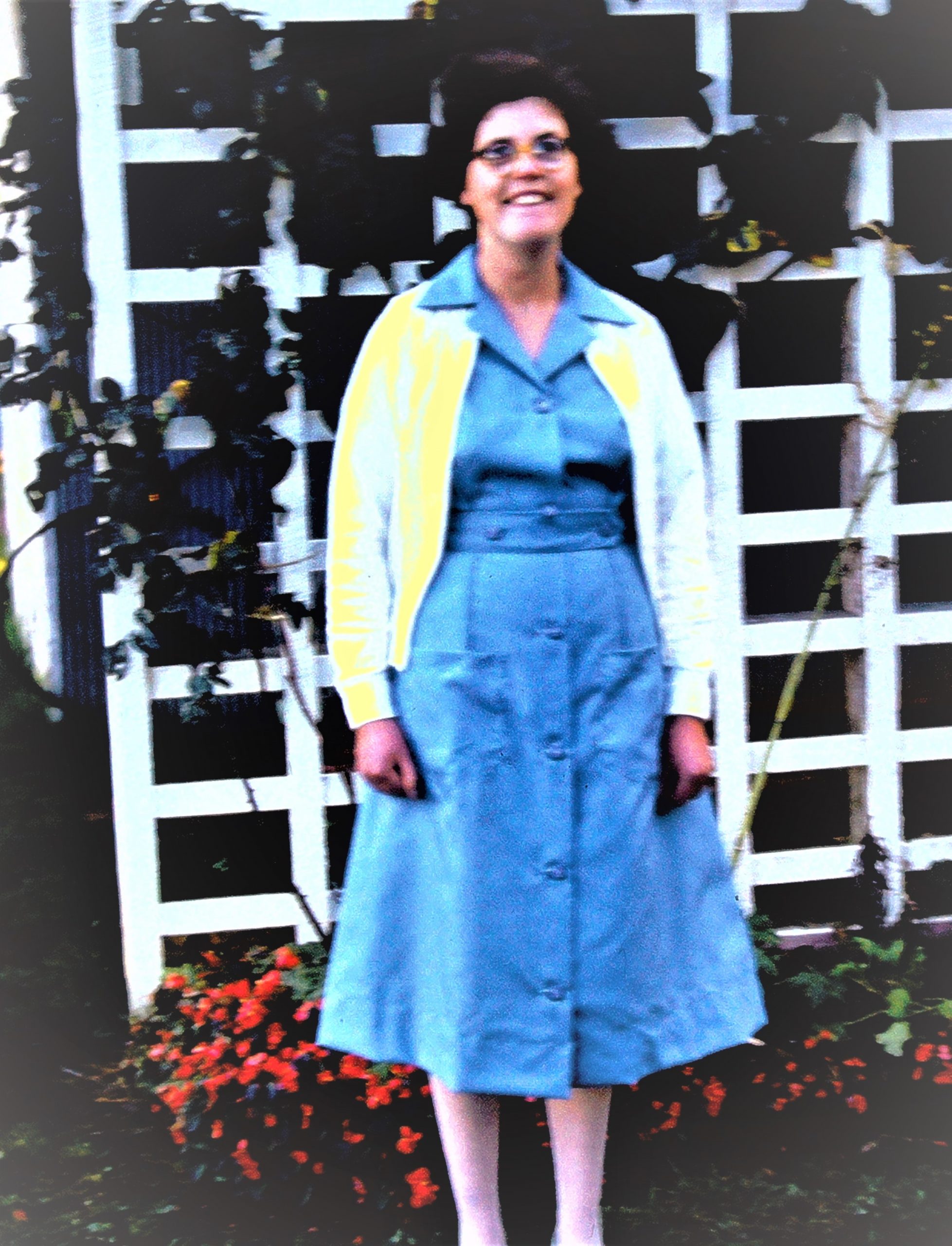 Lesley, smiling, stands in front of a garden trellis. She wears a blue uniform dress with white cardigan