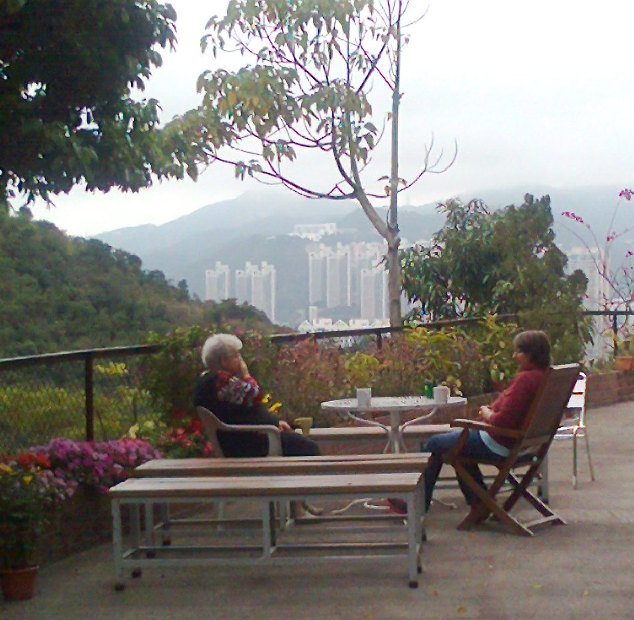 Lesley and another woman are seated facing each other on a terrace. Coffee mugs are on the table between them. In the background are the hills and skyscrapers of Hong Kong. Colourful flowers in planter boxes grow beside a fence