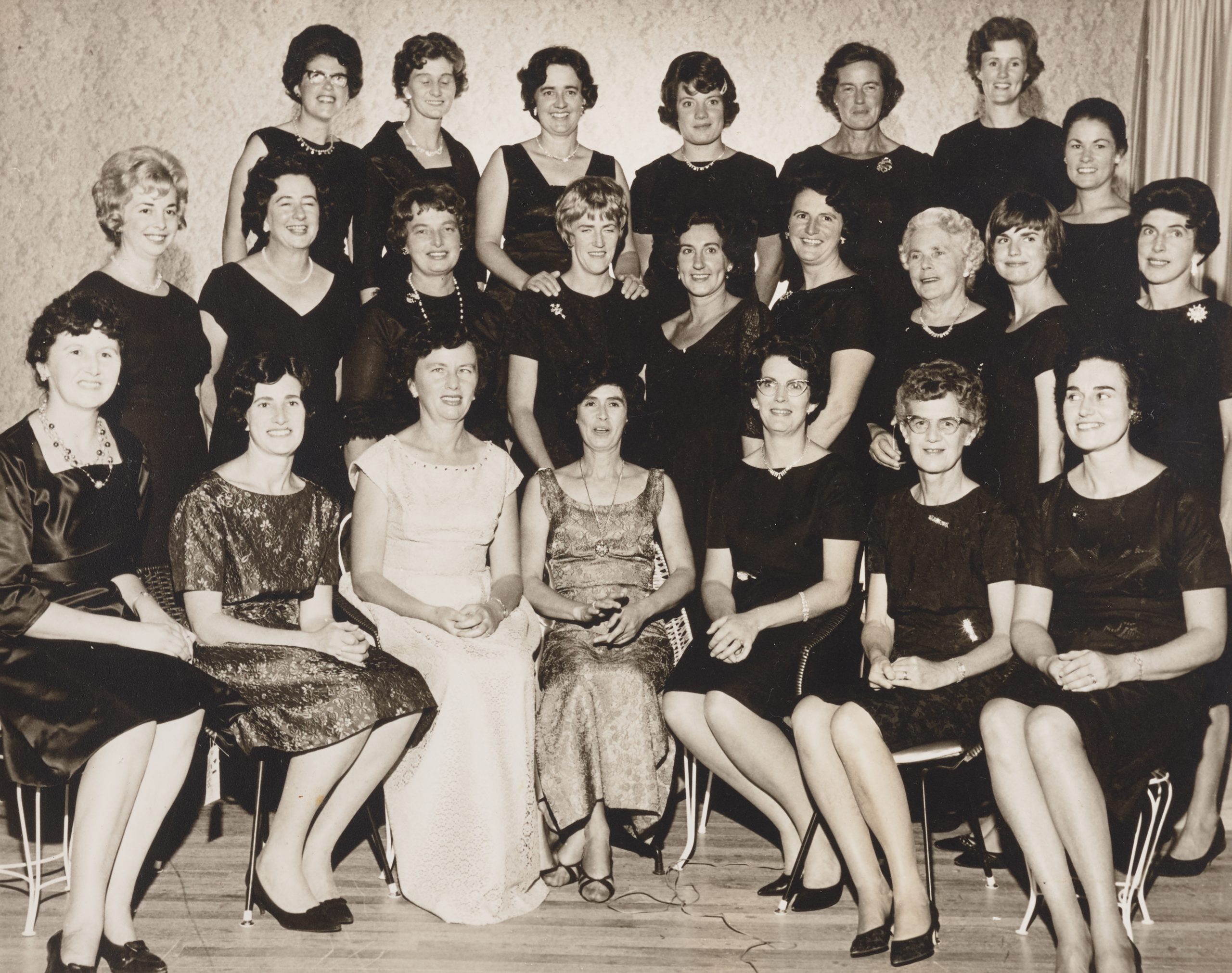 Twenty-three women in three rows are posing for a formal photograph. The conductor and accompanist are seated in the front row. Lesley, standing on a riser in the back row is wearing a sleeveless black dress with a necklace