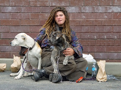 Homeless person sitting on ground with two pet dogs, surrounded by water bottles and paper bags.