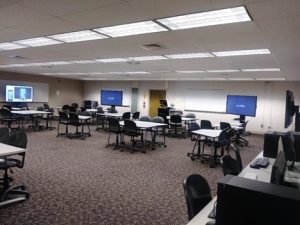 A classroom with a flexible open seating arrangement. Moveable desks and chairs enable students to work in groups while turning furniture to be able to see the multiple projector screens spaced throughout the classroom.