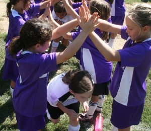 A photo of young girls dressed in soccer uniforms forming a tunnel with their hands for which other girls run through as a post-game ritual.