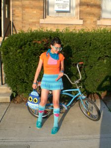 A young woman in brightly colored clothes are carrying an owl handbag is shown standing in front of a vintage blue bicycle, a large hedge, and a town house.