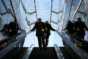 A man and a woman, both wearing business suits, are shown from behind at the top of an escalator.