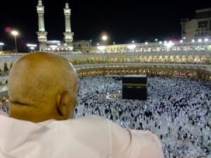 A man dressed in white is shown from behind looking down over the Kaaba, Islam's most sacred site. Hundreds of other people, dressed in all black or all white, can be seen circling a large black cube-like structure on the floor of a stadium-like structure.