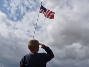 A young boy is shown from behind saluting the American Flag flying from a flagpole