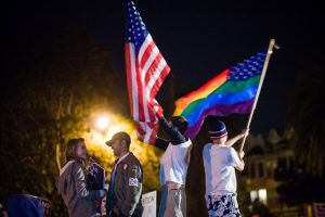 Figure (c) shows people waving a U.S. flag and a rainbow flag