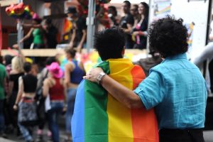 Two people standing with their backs to the camera. One person has a rainbow flag draped over them and the other person has their left arm wrapped around the first person. Multiple pride marchers are visible in the background.