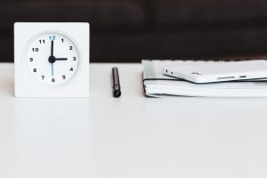 A white clock, black pen, white notebook, and white mobile phone resting on a white desk.