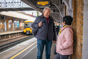 A man and a woman stand together smiling while waiting for a train.